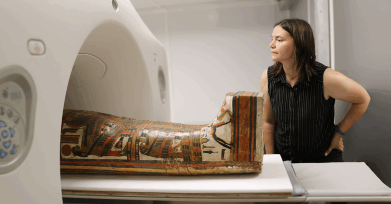 A close-up horizontal shot shows a mummified body in a painted wooden coffin. The coffin is being moved into the opening of a large, modern CT scanner. A person in a striped top and black pants stands to the right, looking on with their hand on their hip. The mummy is identified as Chenet-aa, and the person is a Field Museum staff member. The image captures the intersection of ancient history and modern medical technology.