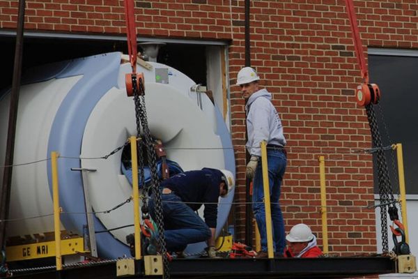 Workers securing a heavy GE MRI machine for a crane lift into a hospital or clinic through a large wall opening.