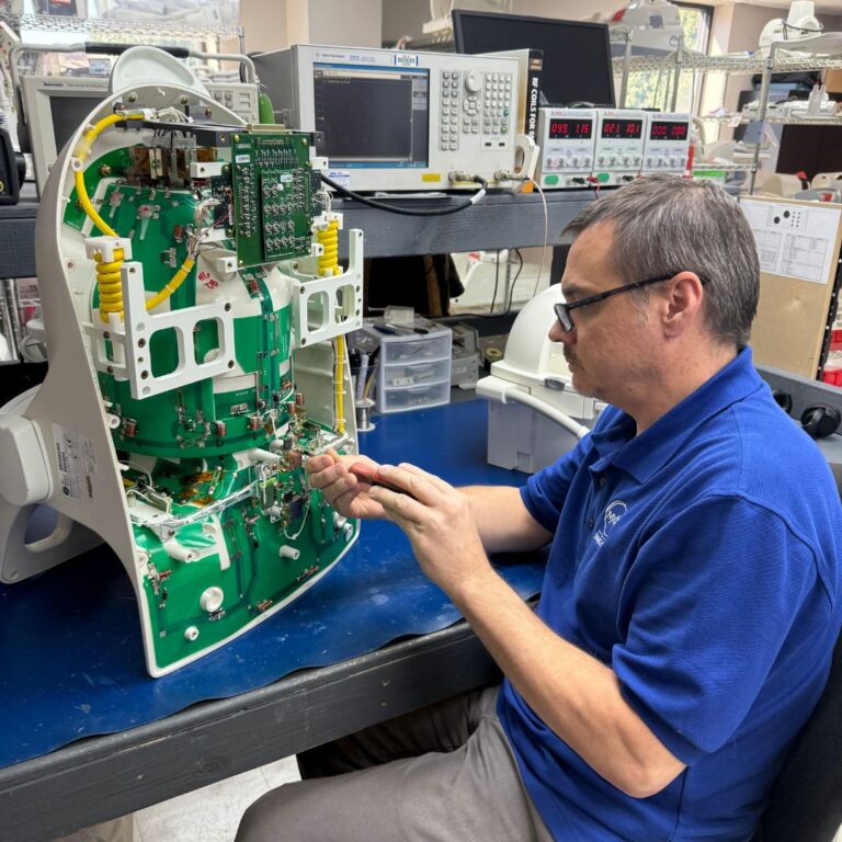 Technician wearing glasses and a blue MXR polo shirt repairs the exposed green circuit board inside a piece of a medical device.