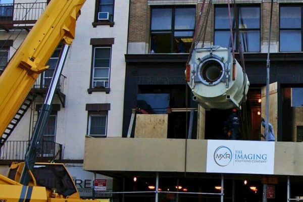 Heavy equipment installation of a medical scanner being craned into a facility over temporary plywood barriers and signage.