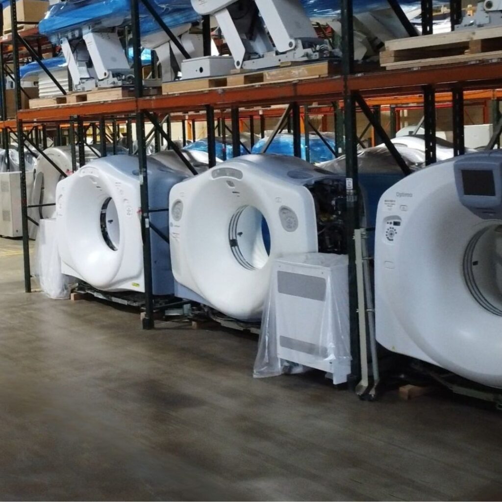 Row of large, white medical scanners (CT or MRI machines) with plastic wrapping stored on a warehouse floor for inventory.